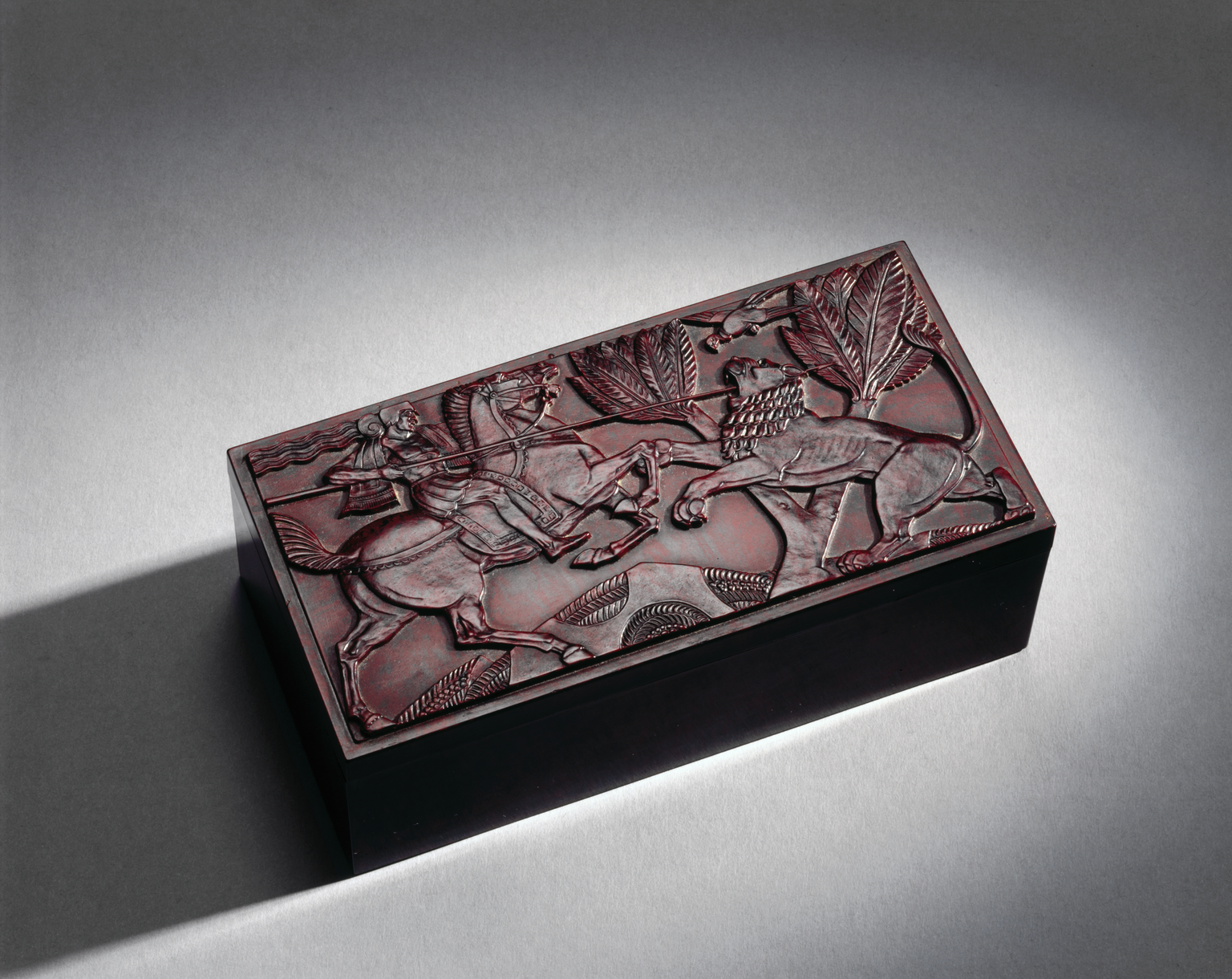 Rectangular cigarette box of imitiation walnut phenolic (Bakelite). Hinged lid decorated with moulded horseman spearing a lion in the Assyrian style, made by Birkby's Ltd., England, 1930s, under trade name "Elo Ware".