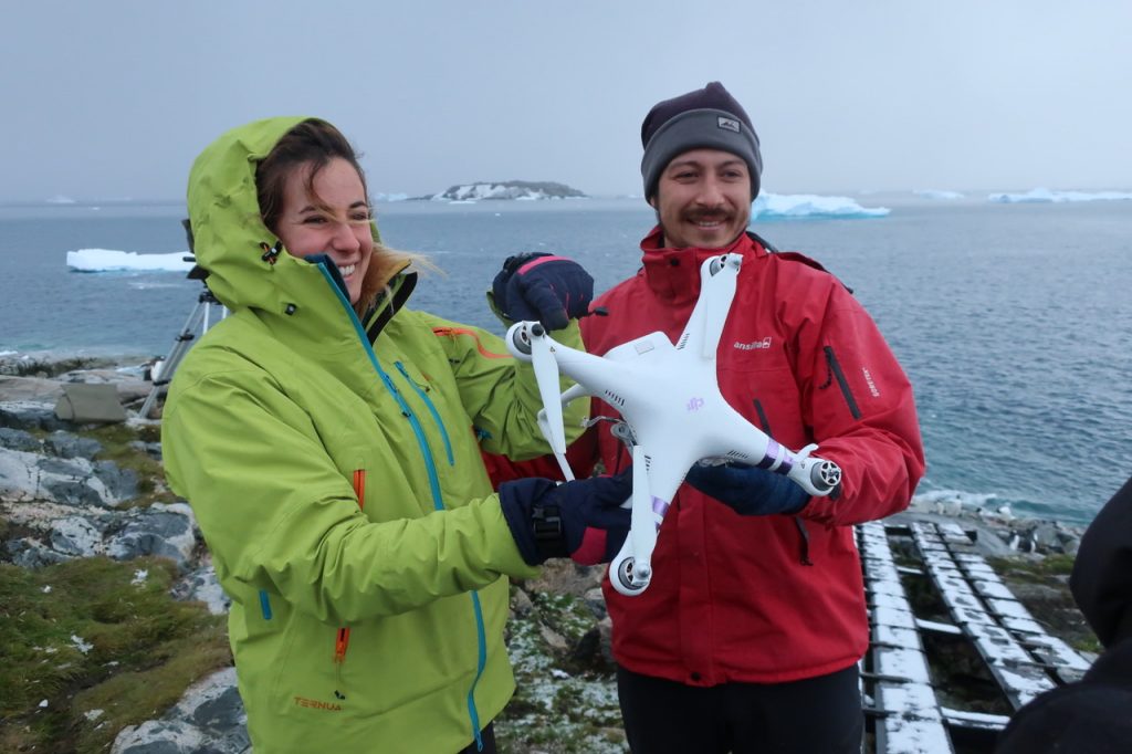 Two scientists stand on the coast of Antarctica holding a drone and smiling to camera.