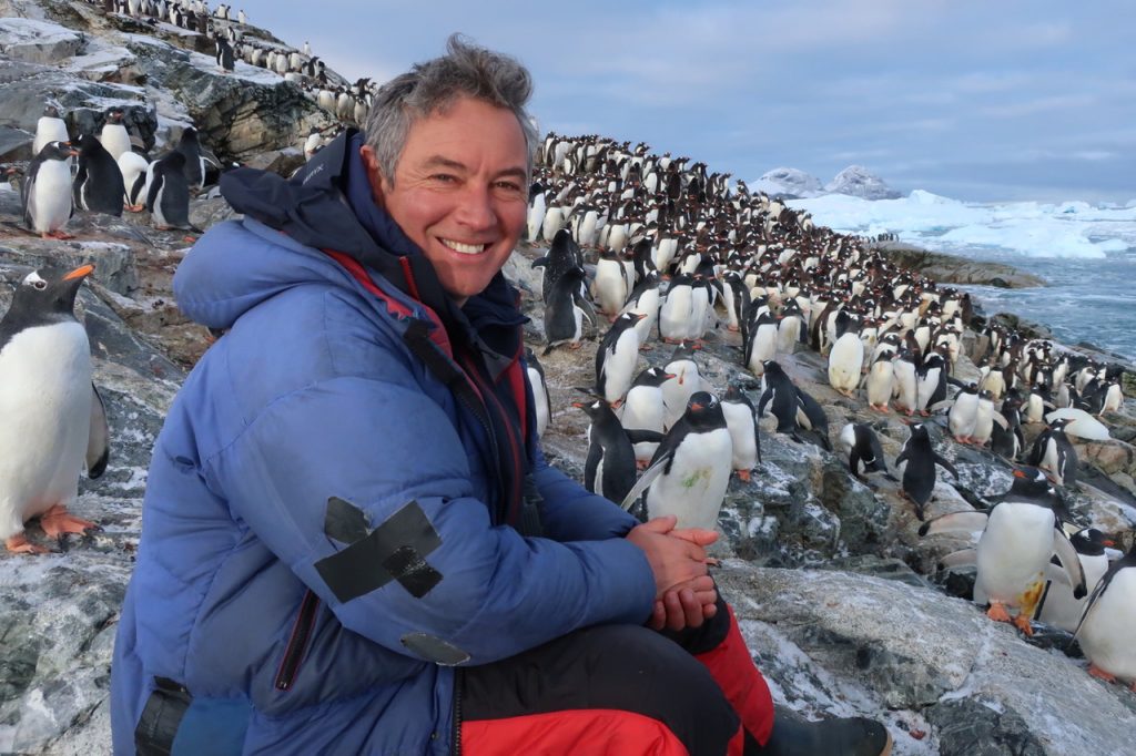 Executive Producer Jonny Keeling sitting on the Antarctic coastline surrounded by thousands of penguins.