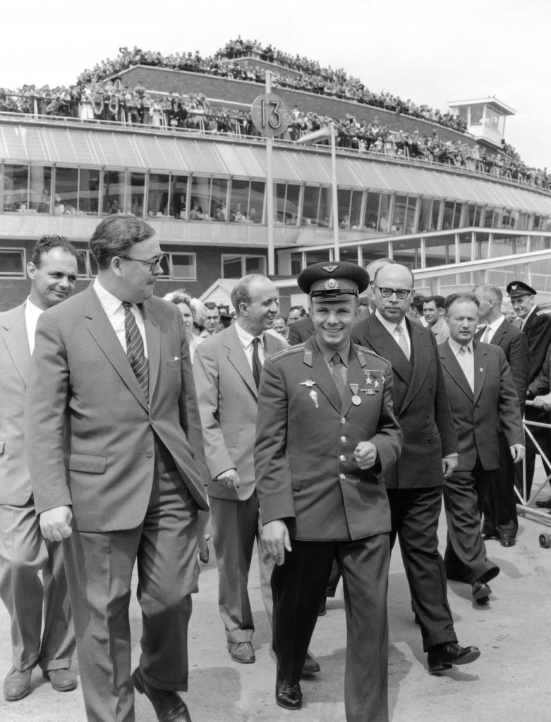 A uniformed Yuri Gagarin at Heathrow Airport followed by a crowd of people. Large crowds of people can be seen filling the roof, balconies and windows of the building in the background.