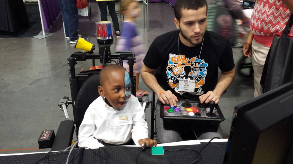 A young boy in a wheelchair watching a man play with a gaming controller.