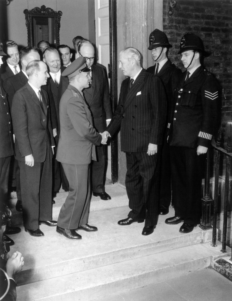 A uniformed Yuri Gagarin shakes hands with then Prime Minister Harold MacMillan on the front steps of a building while crowds and police officers observe from the doorway.