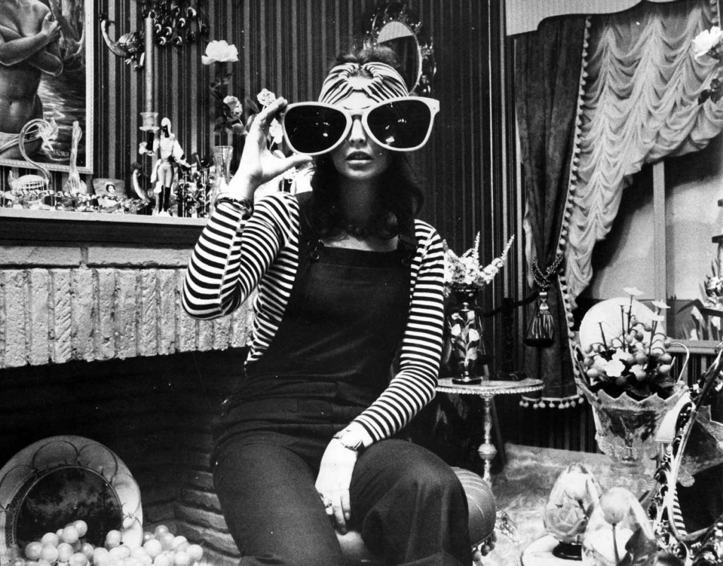 A woman poses with oversized novelty sunglasses in a room filled with knick-knacks